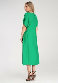 Grünes, knielanges Kleid mit kurzen Ärmeln und seitlichen Raffungselementen. Glatte Textur, lässige Passform und hervorgehobene Öffnung am Rücken in Form eines Schlüssellochs.