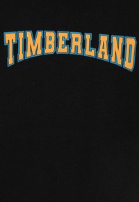 Černá tkanina s nápisem "TIMBERLAND" tučným žlutým písmem orámovaným modrou barvou, uspořádaným v zakřiveném tvaru.