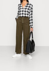 Chemise à carreaux noir et blanc à manches longues, pantalon ample vert olive avec cordon de serrage, baskets blanches et sac en cuir noir.