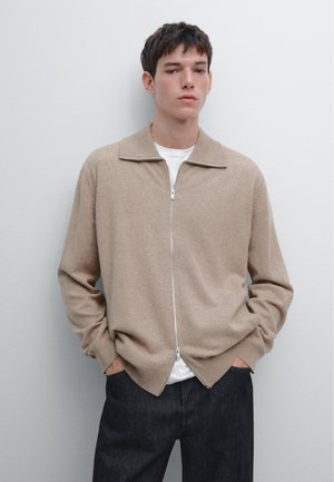 Junger Mann steht an einer weißen Wand, trägt einen beigen Reißverschluss-Pullover über einem weißen Hemd und dunklen Jeans, mit den Händen in den Taschen.