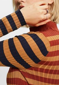 Femme portant un pull à col roulé côtelé avec de larges rayures horizontales en rouille, moutarde et marine, main près du visage avec des bagues argentées aux doigts.