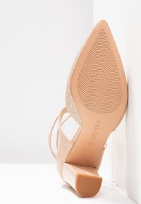 Chaussure nude à talon haut avec un bout pointu et un accent texturé doré sur le talon. La semelle est lisse avec une traction striée.