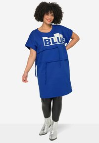 Blauwe oversized jurk met de tekst "BLUE" in het wit. Heeft korte mouwen, een ronde halslijn en een voorzak. Gecombineerd met zwarte leggings en zilveren laarzen.