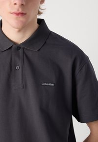 Tmavě šedé polo tričko s knoflíkovým límečkem, krátkými rukávy a malým vyšitým logem Calvin Klein na hrudi. Texturovaná tkanina.