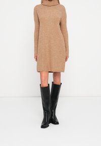 Abito a maglia beige a coste con alto collo alto, maniche lunghe e spacchi laterali, abbinato a stivali alti fino al ginocchio in pelle nera.