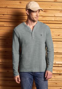 Pitkähihaisen henley-paidan pehmeä, teksturoitu vaaleanharmaa kangas, jossa on kolme nappia kauluksessa ja pieni logo rinnassa. Yhdistetty farkkuihin.