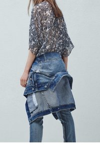 Blusa trasparente a motivi paisley in bianco e nero abbinata a una giacca di jeans stratificata, con diverse tonalità di blu e dettagli logori.