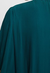 Teal pleatattu kangas, jossa on pystyribbaus, esittelee virtaavaa muotoilua. Kaula-aukko on pyöreä, ja tekstuuri vaikuttaa sileältä.