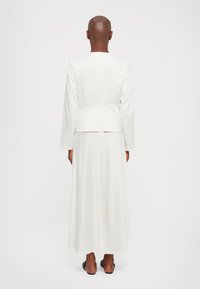 Person mit rasiertem Kopf, der ein langes weißes Kleid und eine maßgeschneiderte weiße Jacke trägt, von hinten gegen einen schlichten weißen Hintergrund betrachtet.