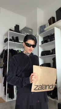 Melnā zip-up kapučedera ar mīkstu tekstūru, turēta abās rokās. Priekšplānā ir brūna papīra soma ar "Zalando" logo, bet fona plaukti ir rastrā.