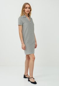 Donna con capelli lunghi biondi, che indossa un vestito a righe a maniche corte e scarpe nere, in piedi davanti a uno sfondo bianco uniforme.