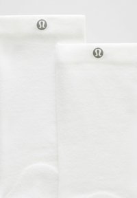 Balti sportiniai kojinės, pagamintos iš tekstūruoto audinio, su patogiu kraštu ir mažai pilkai logotipu kiekvienos kojinės viršuje.