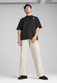 Sort t-shirt med korte ærmer og lodrette striber samt patch-logo, parret med lyse beige brede bukser og sorte sneakers.