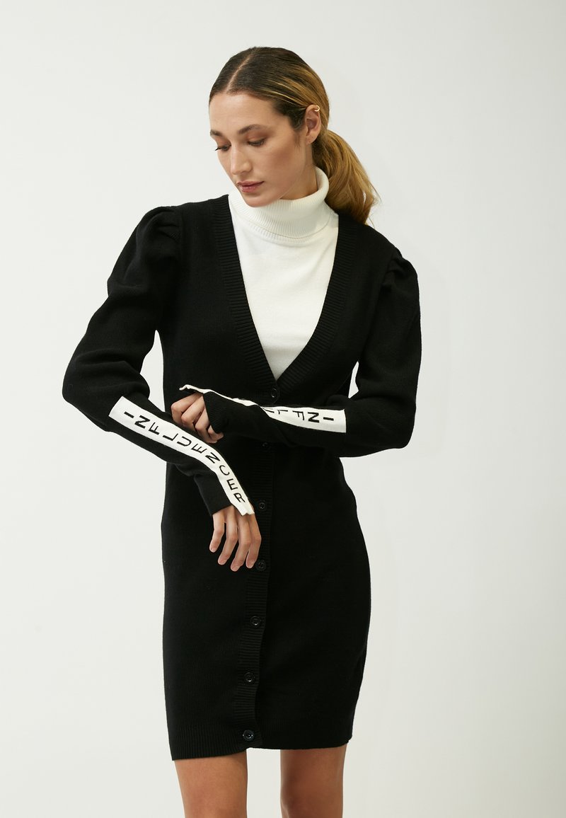 Schwarze gestrickte Strickjacke mit V-Ausschnitt und Knopfverschluss. Merkmale sind gepuffte Ärmel und weißer Text an den Bündchen. Darüber getragen wird ein weißer Rollkragenpullover.