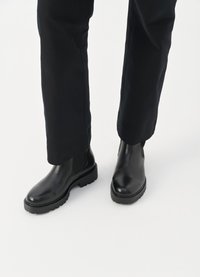 Črne usnjene Chelsea čevlje z elastičnimi stranskimi paneli in trdnimi podplati, kombinirane s črnimi rahlo zoženimi hlačami na beli podlagi.