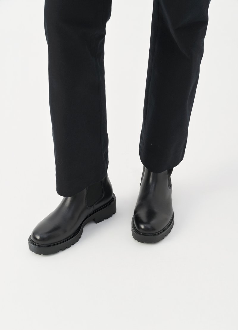 Sorte læder Chelsea-støvler med elastiske sidelameller og robuste såler, båret med sorte let tilspidsede bukser på en hvid baggrund.