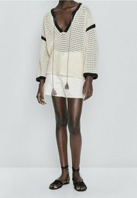 Pull crème oversize avec des accents noirs, décolleté en V et détails à frange. Associé à une mini-jupe blanche et des sandales à lanières noires.