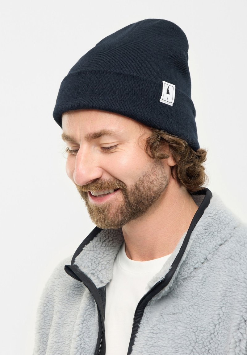 Uomo con capelli ricci e barba che indossa un berretto blu navy con un logo ad albero e una giacca in pile grigia chiara, sorridendo con gli occhi chiusi.