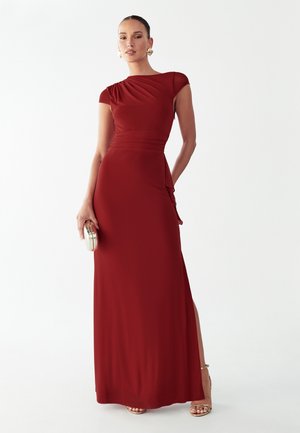 Vrouw draagt een donkerrode, nauwsluitende jurk tot op de grond met korte mouwen, houdt een kleine metallic clutch vast en draagt gouden sandalen en oorbellen.