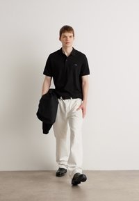 Sort polo skjorte, hvide bukser og sorte sko. Model holder en sort jakke. Enkelt design, glat tekstur, neutral farvepalette.