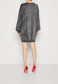 Abito oversize nero scintillante con maniche lunghe, polsini a coste e orlo arrotondato, abbinato a scarpe con tacco rosso lucido.