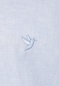 Tela de color azul claro con una textura suave, que presenta un pequeño colibrí bordado en un azul ligeramente más oscuro, centrado en la prenda.