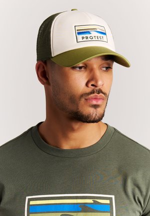 Man draagt een olijfgroene shirt en een bijpassende groen-witte pet met het logo "PROTEST" en een golfpatroon, kijkt naar de zijkant.