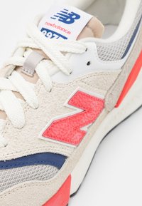 Šedé semišové tenisky s bílou síťovinou, s červeným gumovým logem "N", bílými tkaničkami a modrým akcentem poblíž podešve.