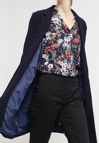 Marineblazer over en blomstret bluse med mørke farver. Blusen har klare blomster; bukserne er sorte og glatte i strukturen.
