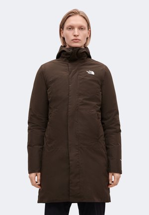 Un mannequin se tient face à l’objectif, portant un long manteau North Face brun foncé à col montant, sur un fond clair uni.