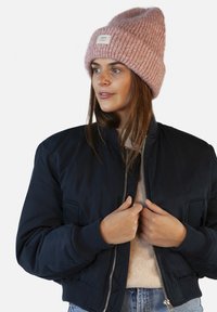 Giacca bomber navale con polsini a costine, dotata di tasche laterali e zip argentata. Abbinata a un berretto lavorato a maglia rosa chiaro.
