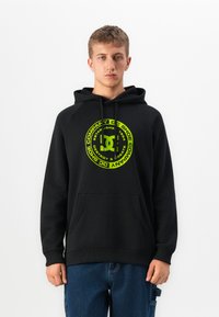 Young man with short hair wears black hoodie with neon green circular logo and blue jeans, standing against plain white background.