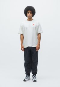 Hvid oversized t-shirt med et lille rødt logo, sorte tapered bukser og grå sportsko. Modellen står foran en ensfarvet hvid baggrund.