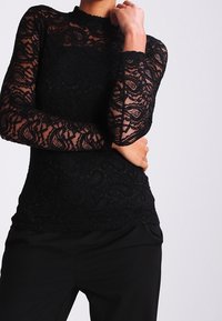 Sort blonde-lace top med lange ærmer, høj hals, blomstermønster, semi-transparent tekstur, lagdelt over en ensfarvet sort base, der komplementerer tætsiddende sorte bukser.