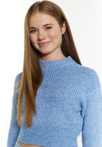 Világoskék, rövidített pulóver bordázott textúrával, magas nyakkal és hosszú ujjal. A szöveten finom színvariációk figyelhetők meg.