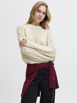 Crème gebreide sweater met kabelpatronen, ronde halslijn en lange mouwen. Gecombineerd met donkere jeans en een kastanjebruine stoffen riem die in de taille is geknoopt.
