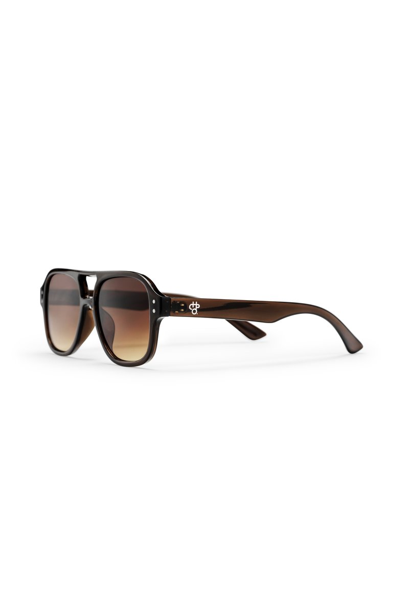 Lunettes de soleil écaille marron avec des verres dégradés marron et un pont en forme de trou de serrure, posées sur une surface blanche face à gauche.