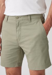 Gros plan d'une personne portant un short vert clair jusqu'aux genoux avec des poches et une chemise blanche rentrée, une main dans une poche.