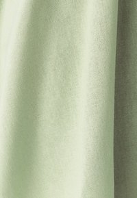 Šviesiai žalia audinių medžiaga su matoma tekstūra ir minkštais raukšlėmis, sukuriančiomis šešėlius ir šviesos atspindžius.