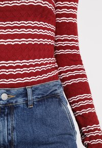 Rode textuur lange mouwtop met witte golvende strepen, gecombineerd met hoog getailleerde blauwe denim jeans met een knoopsluiting en vijf zakken.