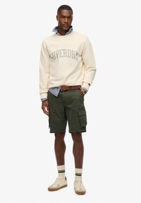Felpa crema con logo "SUPERDRY", pantaloni cargo oliva, sneakers beige e calzini a righe bianchi e verdi, che mostrano uno stile casual.