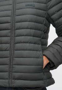 Prim-plan al unei persoane care poartă o jachetă matlasată gri închis, cu fermoar frontal vizibil, cu mâna în buzunarul lateral și logo-ul Calvin Klein pe piept.