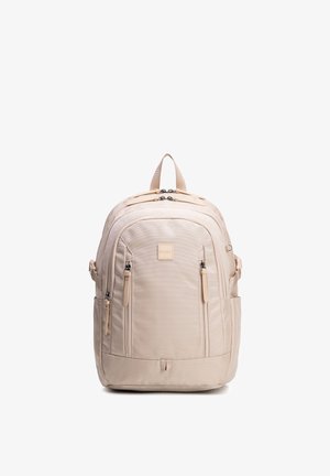 Mochila beige con múltiples compartimentos con cremallera, bolsillos laterales, correas acolchadas y una asa superior, de pie sobre un fondo blanco.