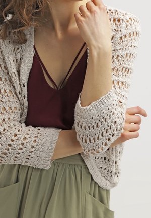 Gilet au crochet de couleur beige clair avec un tissage ouvert, associé à un débardeur bordeaux profond. Modèle portant un pantalon ample vert olive.