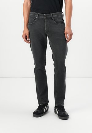 Jeans en denim gris à coupe slim, avec un design à cinq poches, des passants de ceinture et une fermeture éclair avec bouton. Portés avec des baskets noires.