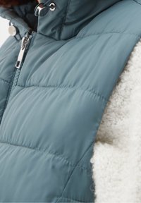 Vadderad väst i teal med hög krage, med dragkedja och metallbeslag. Texturen kontrasterar med det vita, fluffiga innerfodret.