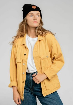 Żółta kurtka z kanwy z dwoma przednimi kieszeniami, srebrnymi zatrzaskami i podwiniętymi rękawami, w zestawie z białym t-shirtem i niebieskimi dżinsami. Czarna beanie z odznaką.