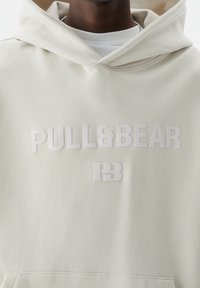 Primo piano di una persona che indossa un hoodie beige chiaro con il logo in rilievo "PULL&BEAR" e "PB" sulla parte anteriore.