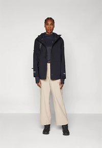 Femme se tenant de face, portant une veste à capuche noire, un t-shirt noir, un pantalon large beige, des bottes noires et un sac à dos noir.