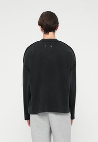 Čierna oversized mikina s dlhými rukávmi, vysokým golierom a minimálnym prešívaním. Obsahuje dva kovové patentky na zadnej strane.